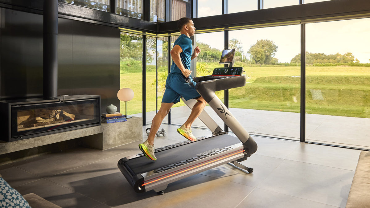 Person using a treadmill in a modern living room with large windows.